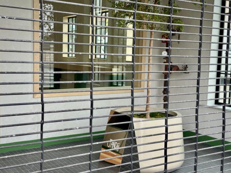 View through metal bars showing a window, a potted tree, and a box labeled "VAND!" in a tiled indoor space.