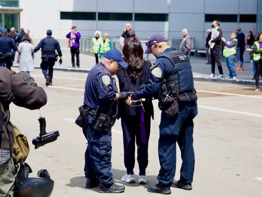 Two police officers arrest a woman with zip ties outdoors, while several people observe in the background.