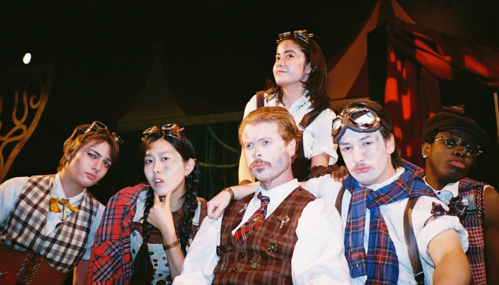 A cast of six actors at UC Berkeley, including Kelly Mou, poses in stylized costumes with a steampunk aesthetic. Several wear goggles on their heads or over their eyes, along with patterned vests, plaid shirts, and newsboy caps, all set against a dark stage backdrop.