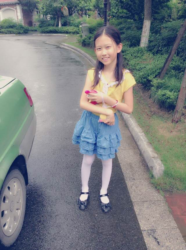 A young Kelly Mou stands on a wet asphalt road next to a green car, wearing a yellow sleeveless top with a white ruffled collar and a tiered blue skirt. She is smiling at the camera and holding a small toy in her hands.