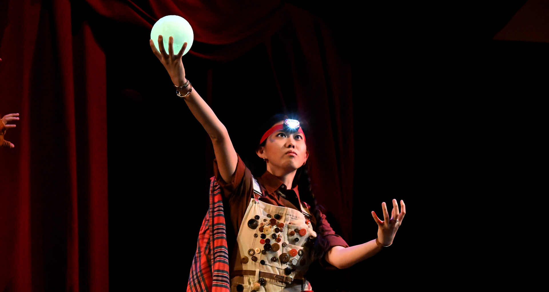 Kelly Mou performs on a dark stage, wearing a headlamp and overalls covered in buttons. She looks upward with an intense, focused expression, holding a glowing green orb high in one hand while her other hand is tensed at her side.