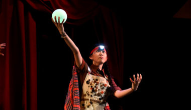 Kelly Mou performs on a dark stage, wearing a headlamp and overalls covered in buttons. She looks upward with an intense, focused expression, holding a glowing green orb high in one hand while her other hand is tensed at her side.