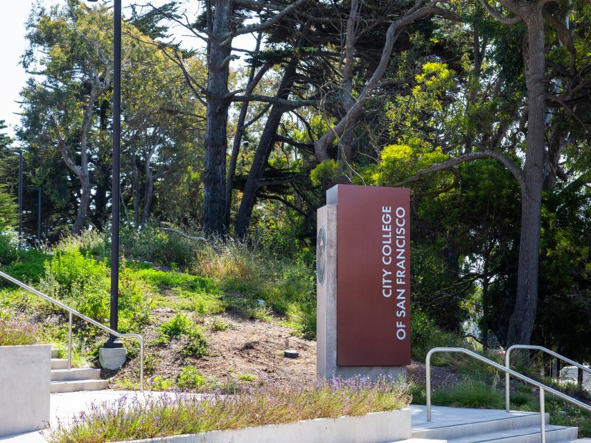 A sign for City College of San Francisco stands next to a path and staircase, surrounded by trees and greenery.
