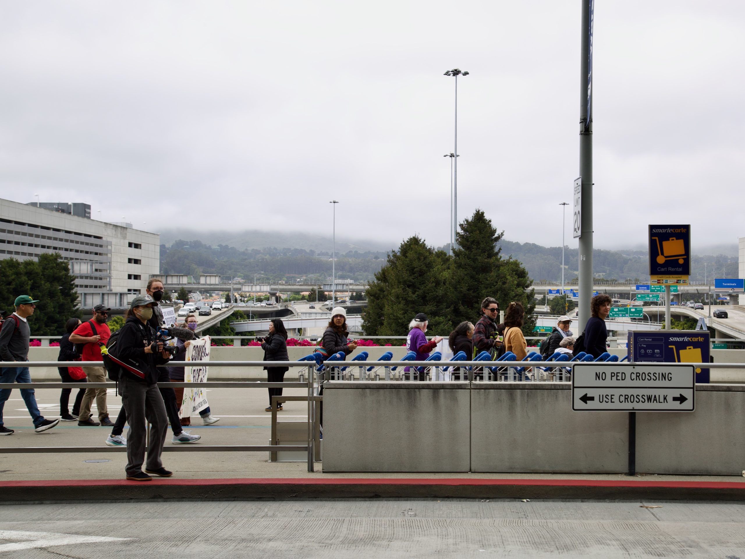 People stand and walk near a row of luggage carts outside an airport terminal, with signs indicating no pedestrian crossing and directions to use the crosswalk to avoid potential arrest for jaywalking.