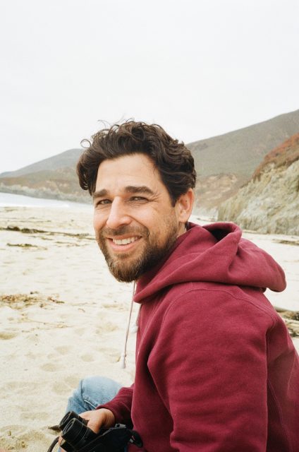 A man with a beard and maroon hoodie sits on a sandy beach, smiling at the camera, with hills and ocean in the background.