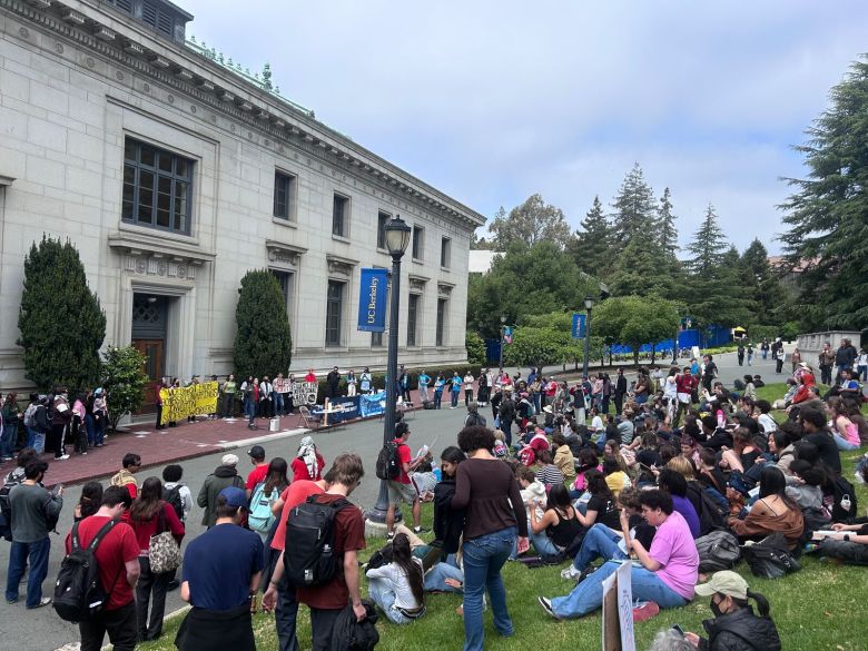 A large group of people sit on a law listening to a speaker in front of an academic building