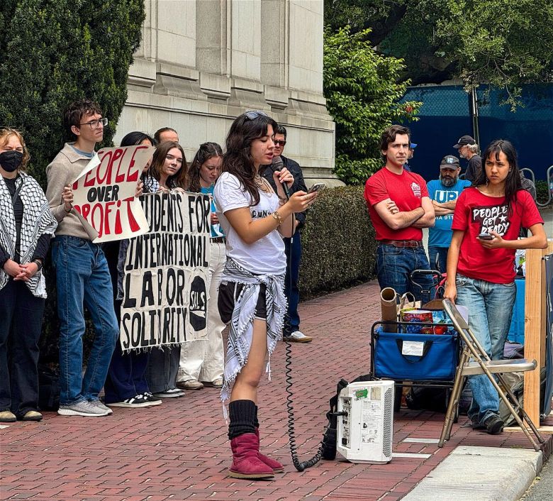 A young person speaks into a microphone, while people behind them hold protest signs