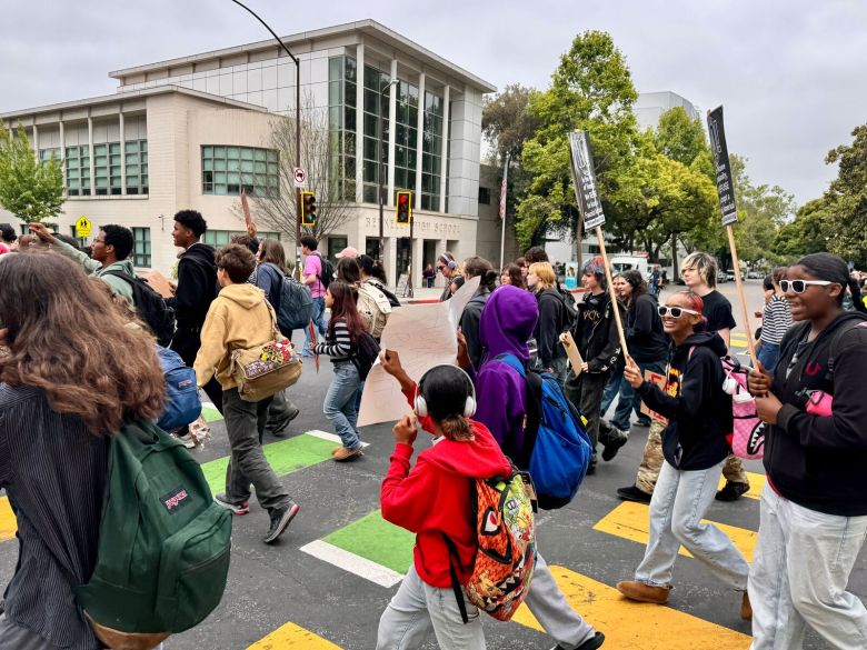A group of high school students, many of them carrying picket signs, march through an intersection in front of Berkeley High School