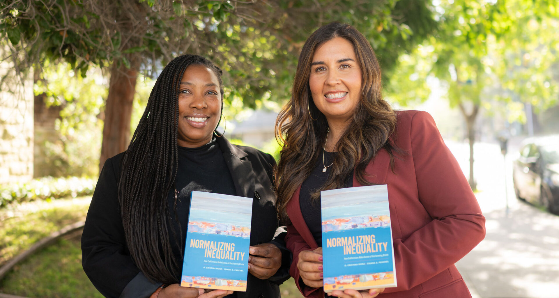 Two women in professional attire, one Black and one Latina, hold up copies of a book with the title Normalizing Inequality.