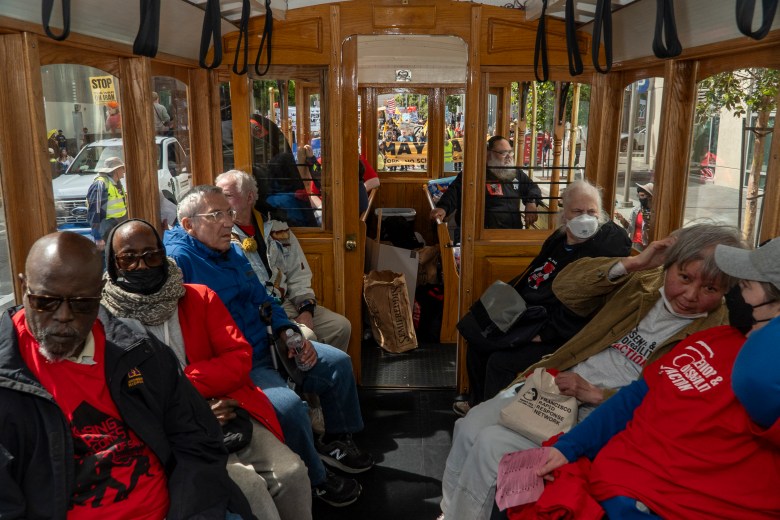 Inside a bus that looks like a cable car interior, people rest while others march outside