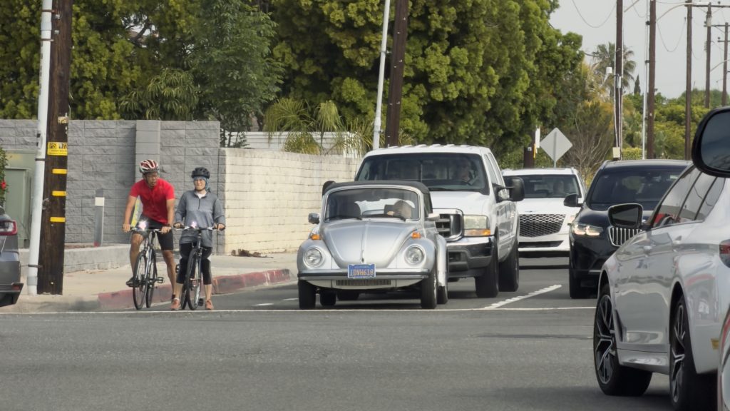 Two cyclists wait to cross a street intersection with many cars