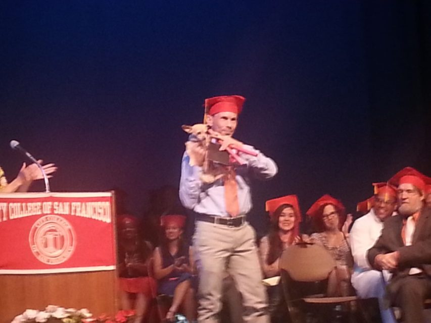 A man in a graduation cap and gown holds a small dog on stage during a graduation ceremony at City College of San Francisco. Several people wearing caps sit in the background.