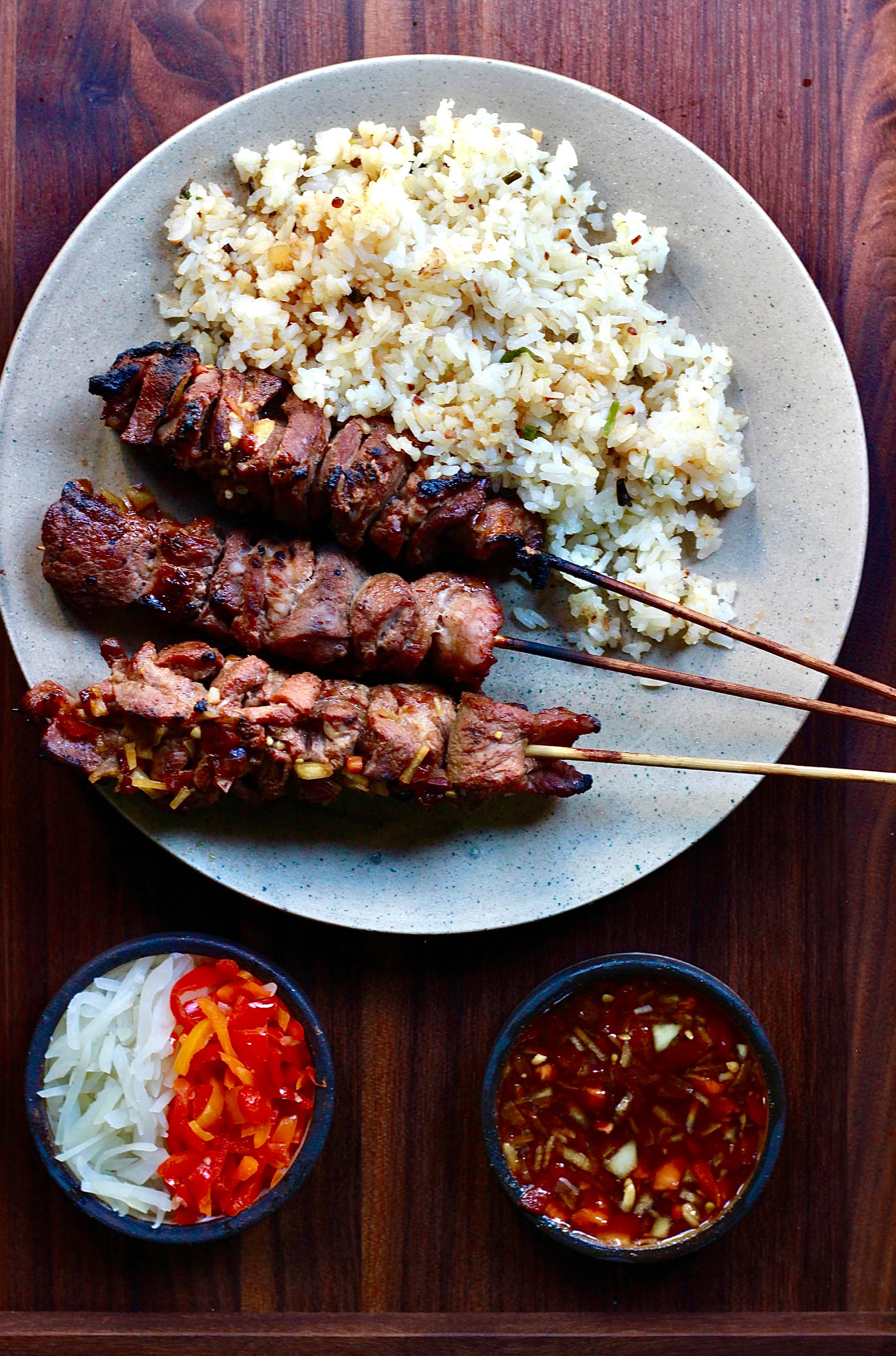 Three skewers of barbecue pork next to garlic rice on a plate, plus two sauces, from Restaurant Naides in San Francisco.