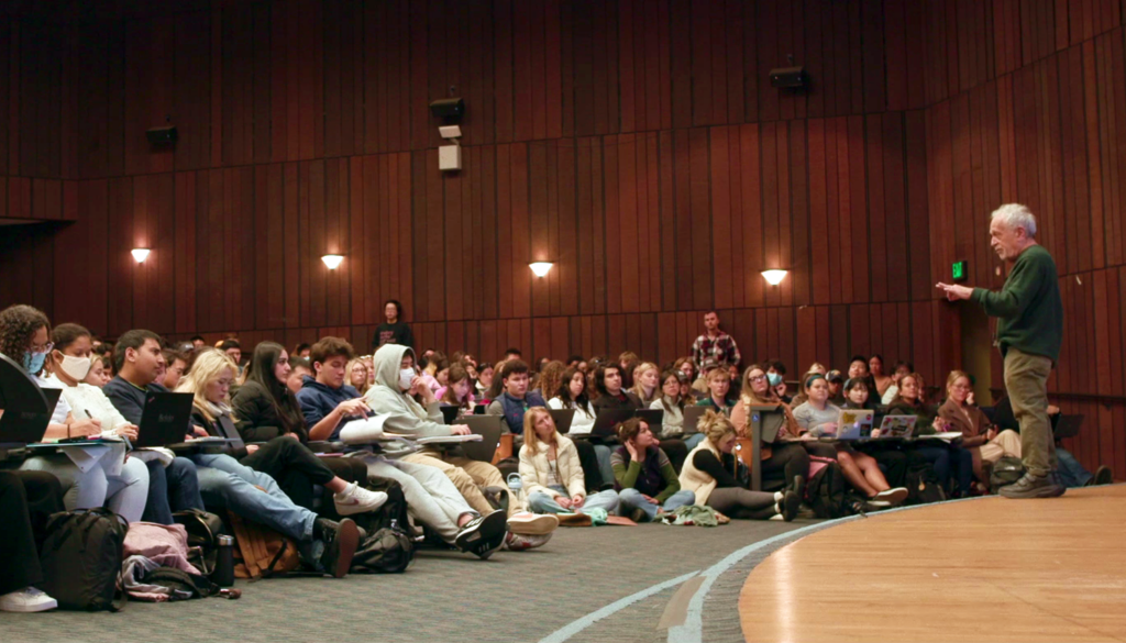 Robert Reich at right, on a speaking platform, with a large auditorium full of students stretching out to the left.