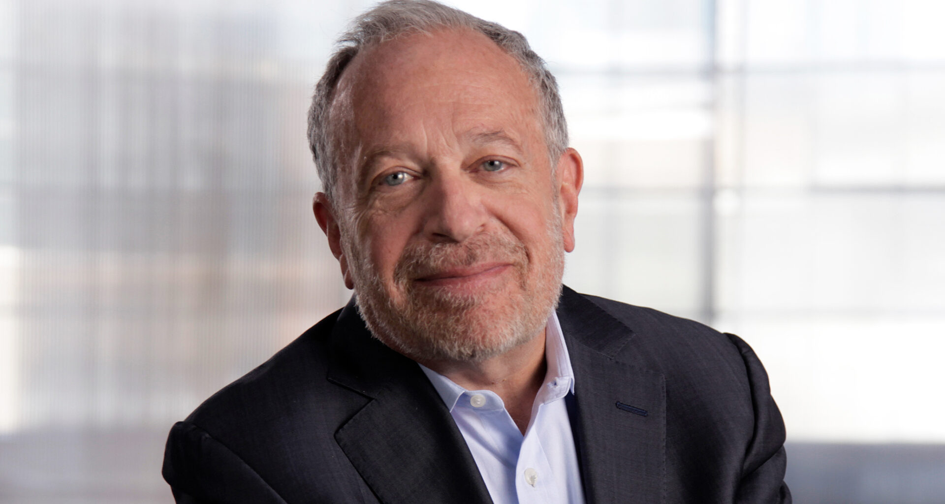 informal horizontal portrait of Robert Reich, wearing a blazer and a light blue shirt, against a bright silver-white background