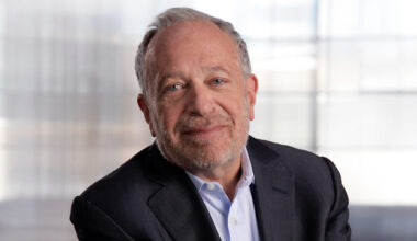 informal horizontal portrait of Robert Reich, wearing a blazer and a light blue shirt, against a bright silver-white background