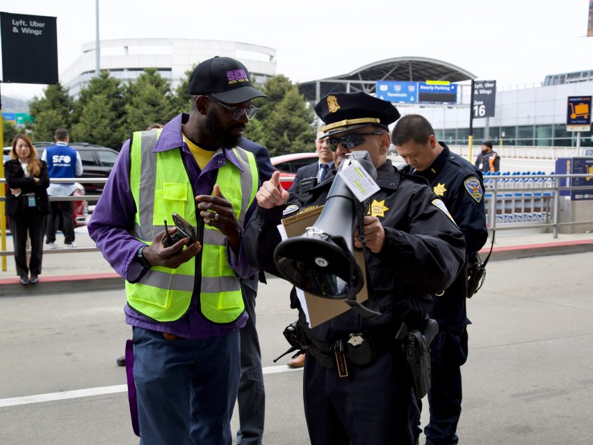 A police officer with a megaphone speaks to a man in a safety vest outside an airport terminal amid an apparent arrest, while other officers and people are visible in the background.