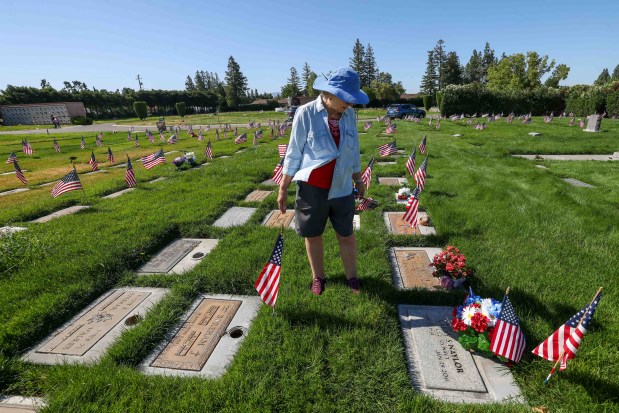 American flags stand in front of the gravestones of fallen soldiers as Conni Naylor stands by the gravestone of her husband, Navy veteran Larry Naylor, at Memory Garden Cemetery in Livermore, Calif., Saturday, May 23, 2026. About 120 mother-and-son teams from the Livermore chapter of the Young Men's Service League, in partnership with members of American Legion Post 47, placed the American flags for Memorial Day weekend at Memory Garden Cemetery and St. Michael's Cemetery. (Ray Chavez/Bay Area News Group)