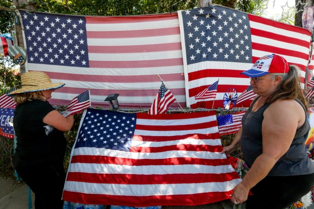Gina Gille, left, and another woman, who only gave a first name of Donna, attaches an American flag as they and others gather for Trump supporter Kerry Sheron, who died on Monday due to injuries he sustained after being beaten last week, while in front of Sheron's home in Escondido on Monday, May 25, 2026. (Hayne Palmour IV / For The San Diego Union-Tribune)
