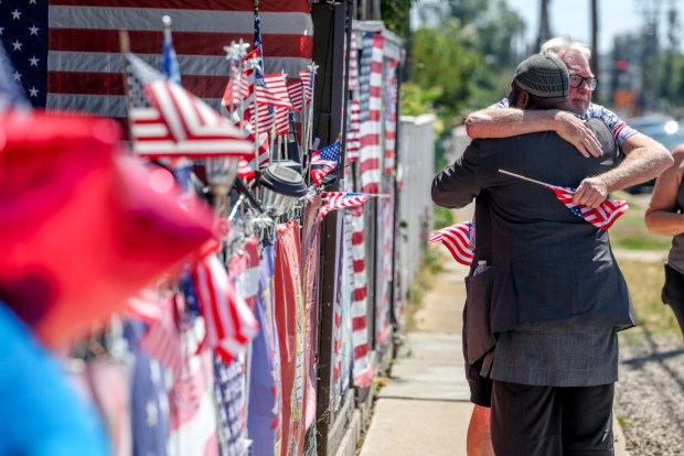 Yusef Miller, front, who said he was there because of being a fellow Escondido resident and veteran, hugs Ron Ammon of Pauma Valley, the father of a veteran, after they worked together to attach flags in front of the home of Trump supporter Kerry Sheron, who died on Monday due to injuries he sustained after being beaten last week, in Escondido on Monday, May 25, 2026. (Hayne Palmour IV / For The San Diego Union-Tribune)