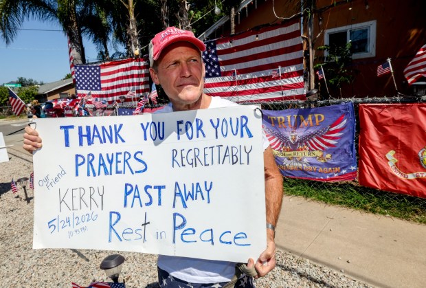 Jim Kelso holds a sign as he stands in front of the home of friend and Trump supporter Kerry Sheron, who died on Monday due to injuries he sustained after being beaten last week, while in front of Sheron's home in Escondido on Monday, May 25, 2026. (Hayne Palmour IV / For The San Diego Union-Tribune)