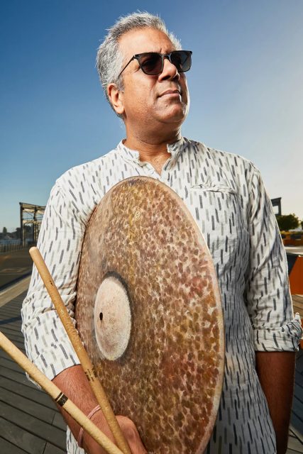 A man in sunglasses holds drumsticks and a large cymbal, standing outdoors on a deck under a clear blue sky.