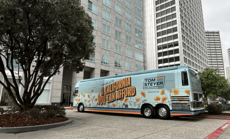 A campaign bus with the slogan "A California You Can Afford" is parked near tall buildings and trees, conveying a political message in an urban setting.
