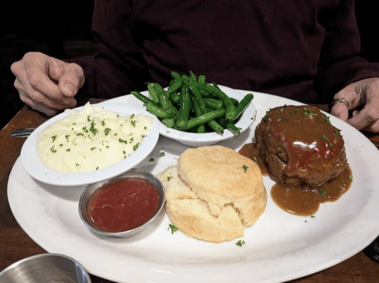 A person sits at a table with a plate of meatloaf with brown gravy, a biscuit, mashed potatoes, green beans, and a side of ketchup.
