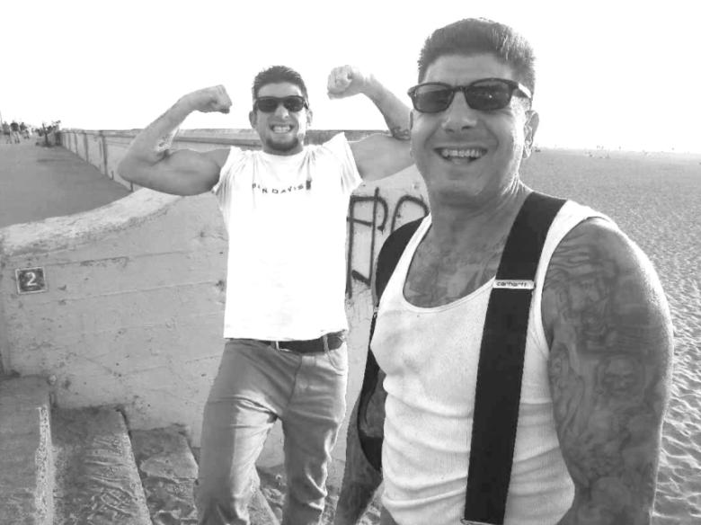 Two men at the beach; one is smiling at the camera in a tank top and suspenders, while the other flexes his muscles and smiles in the background.