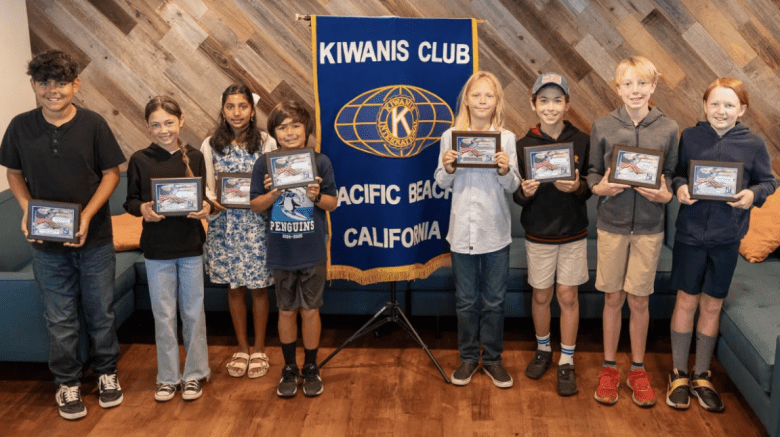 Eight children stand in a row, each holding a framed certificate. They smile proudly in front of a "Kiwanis Club Pacific Beach California" banner.