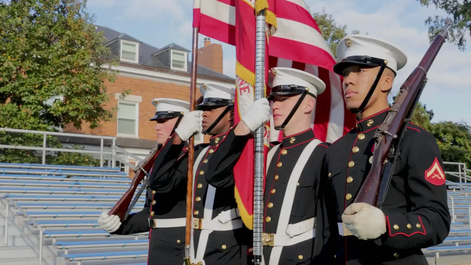 Four Marines in dress blues stand at attention with rifles as they carry the American flag in a color-guard ceremony in a stadium setting.