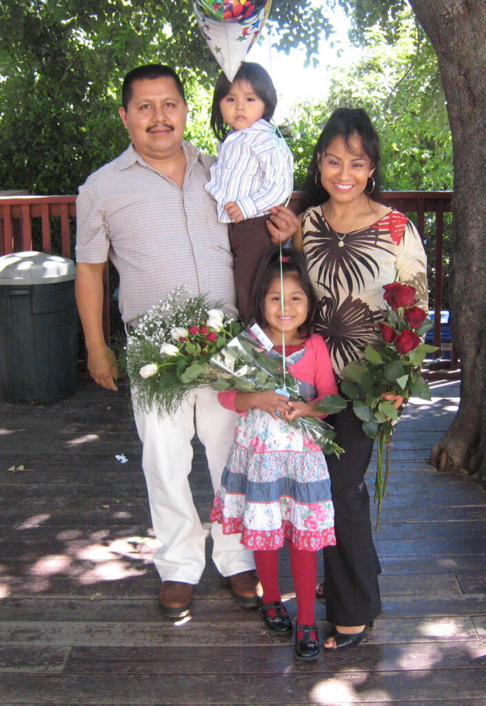 Tania at age 4 poses with her parents and younger brother on a wooden deck outdoors, holding large bouquets of red and white roses and a balloon.