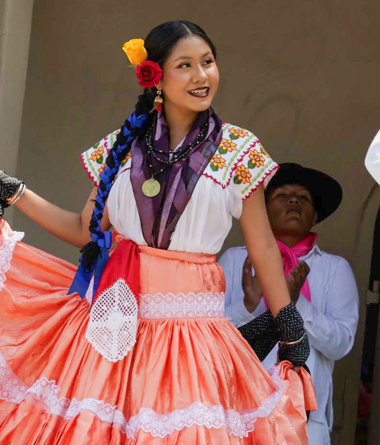 Tania as a teenager performs a traditional folklorico dance in a vibrant orange skirt, an embroidered white blouse and flowers in her hair. 