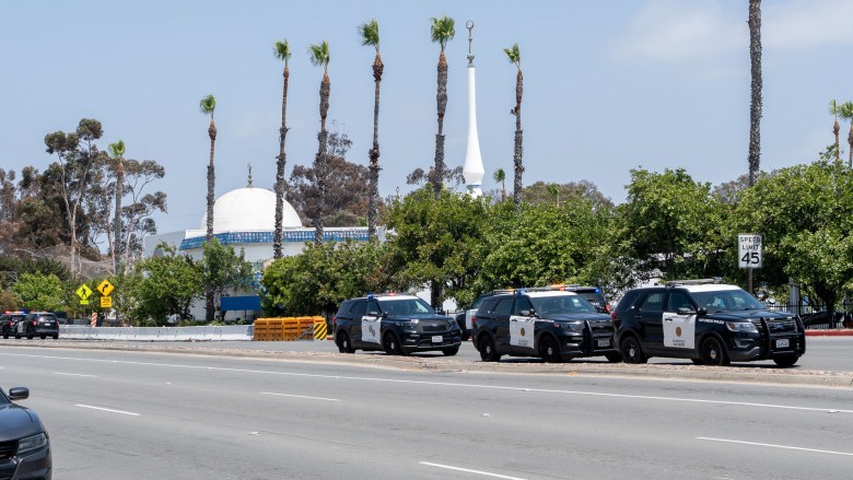 The white dome of a mosque beind palm trees with a wide road and police cars