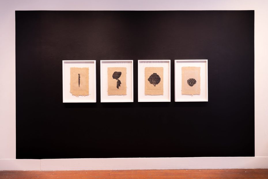 Four framed abstract ink drawings on tan paper are displayed in a row against a black wall in a gallery setting.