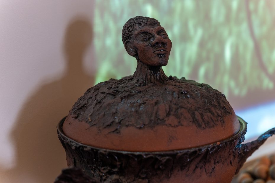 A ceramic pot with a textured lid features a sculpted human head emerging from the top, set against a softly lit background.