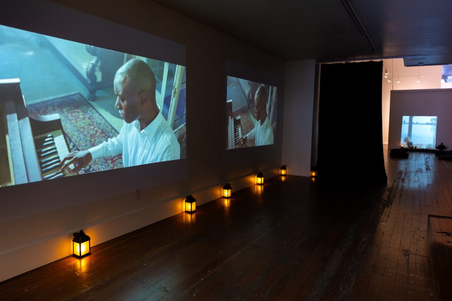 A dimly lit room with video projections of a man playing piano on a wall, lanterns on the floor, and a window in the background.