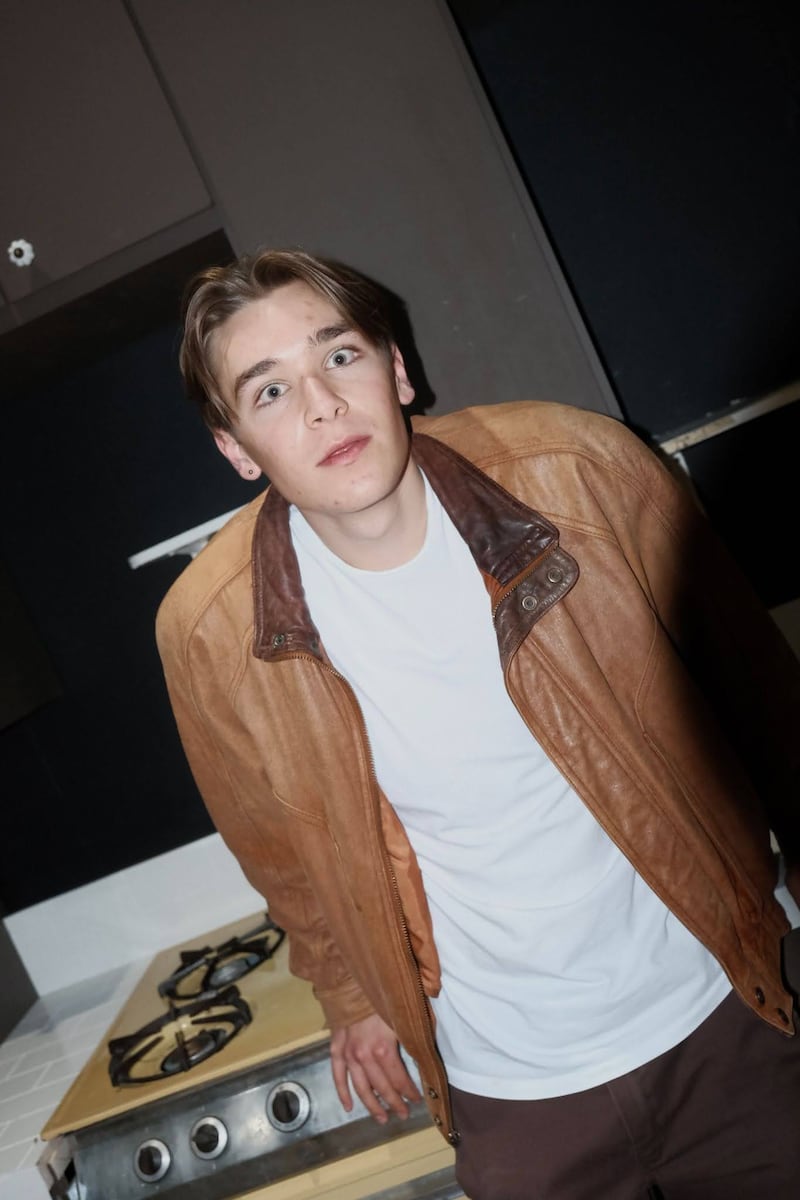 A blonde person in a white t-shirt and brown jacket staring at the camera in the kitchen