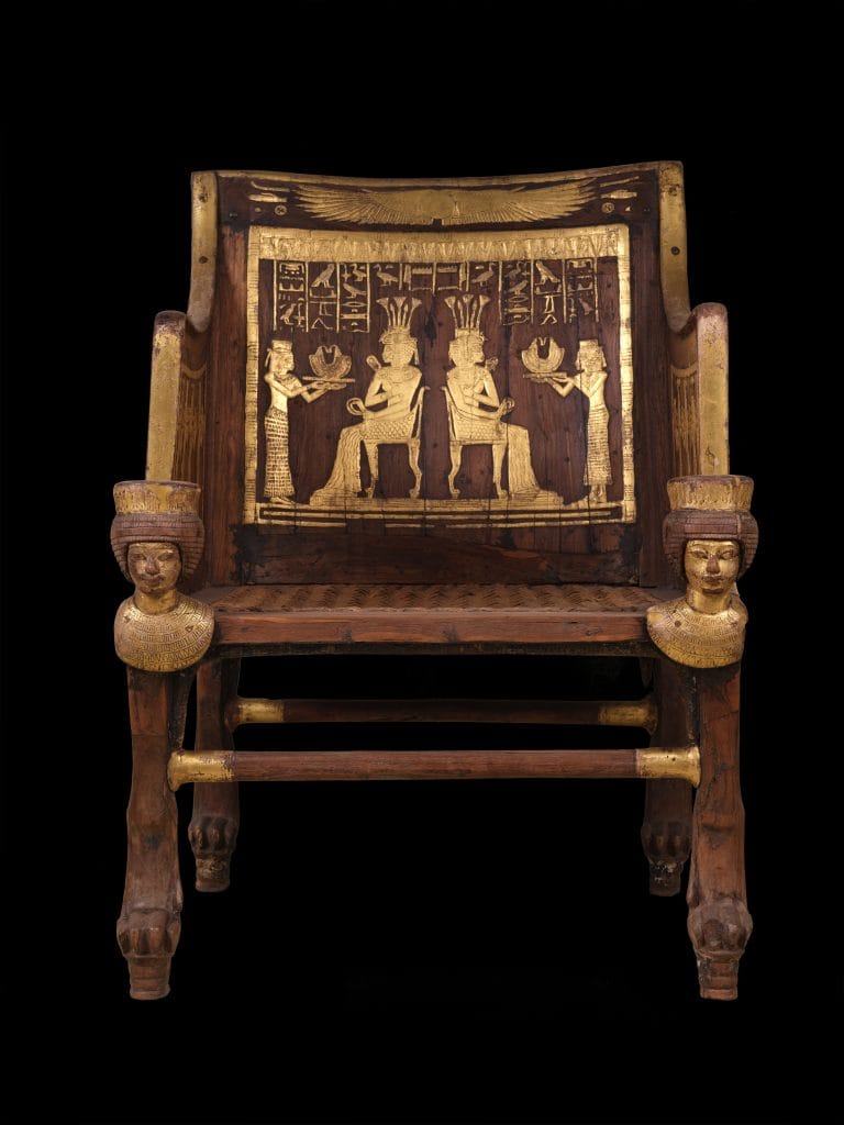 A wooden Ancient Egyptian chair with detailed gold embellishment, including a scene of two figures sitting back to back on it is back—pictured against a totally black background
