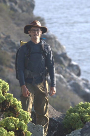 Man in leather hat stands on narrow, rocky path along cliff with ocean in background