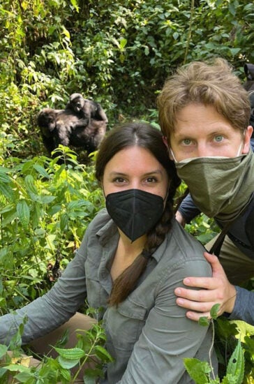 Man and woman wear face masks among jungle bushes as gorilla mother and baby forage behind them