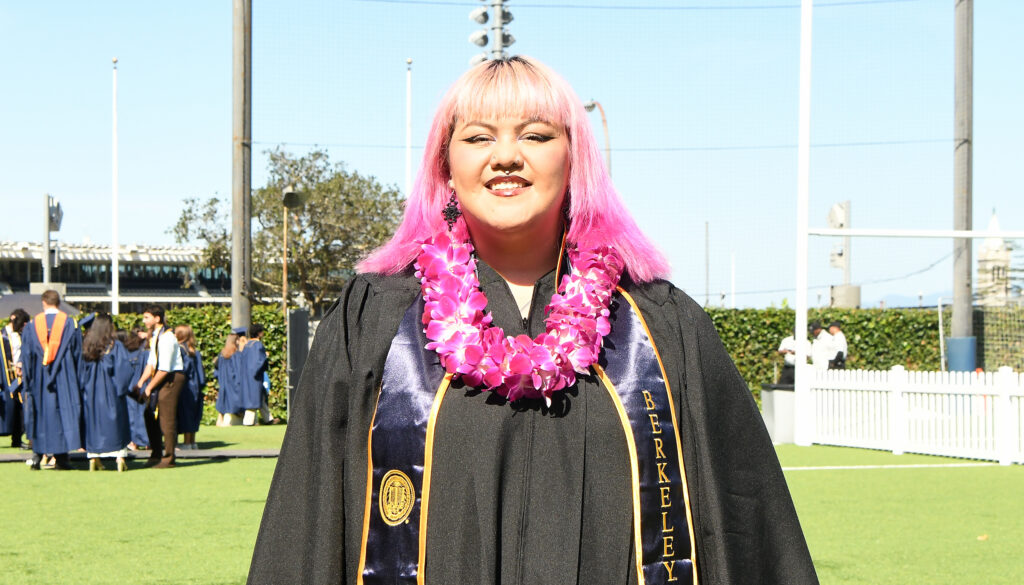A graduate in her robe with a stole, a pink lei, and pink hair smiles at the camera