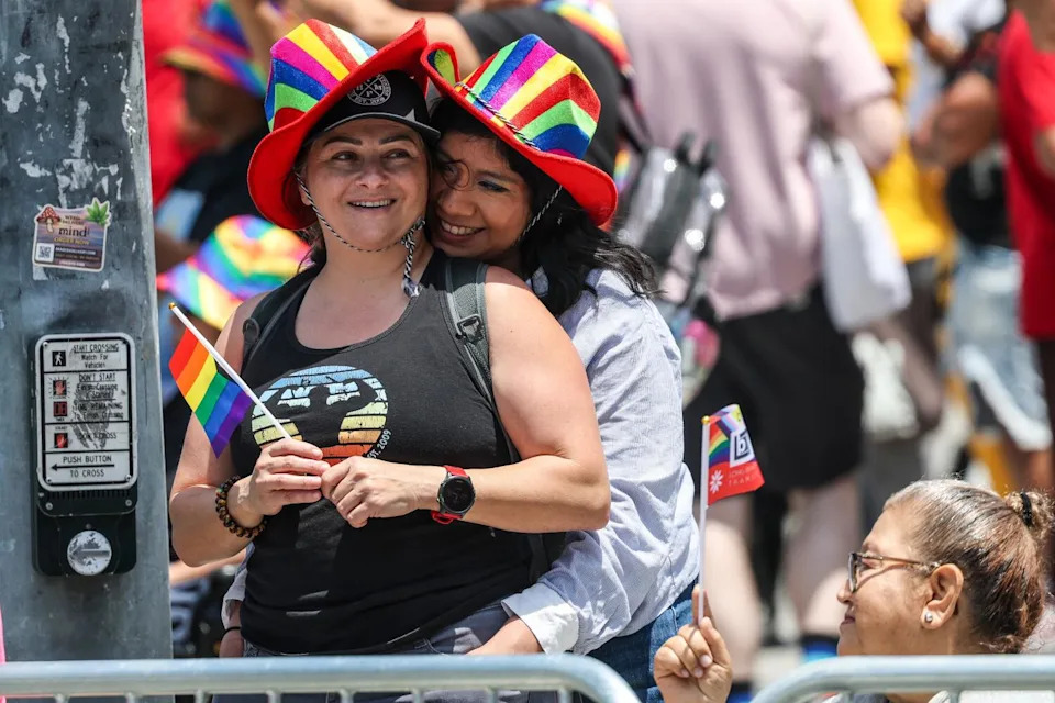 Two people in rainbow hats watch the Pride parade.