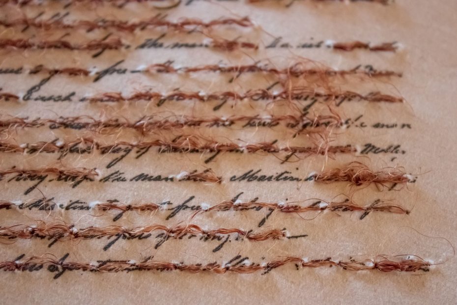 Close-up of handwritten text on paper partially obscured by rows of beige and brown threads sewn across the surface.