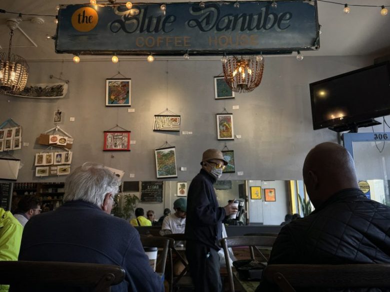 Interior of a coffee house with several people seated, art on walls, and a "Blue Danube Coffee House" sign above. One person stands wearing a hat and mask, and ceiling lights are visible.