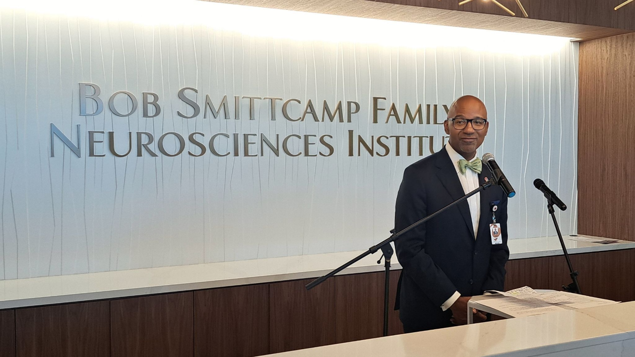 Image of Dr. Ian Johnson, dressed in a dark suit, white shirt, and pastel green bow tie, standing in front of a Bob Smittcamp Family Neorosciences Institute sign inside CRMC