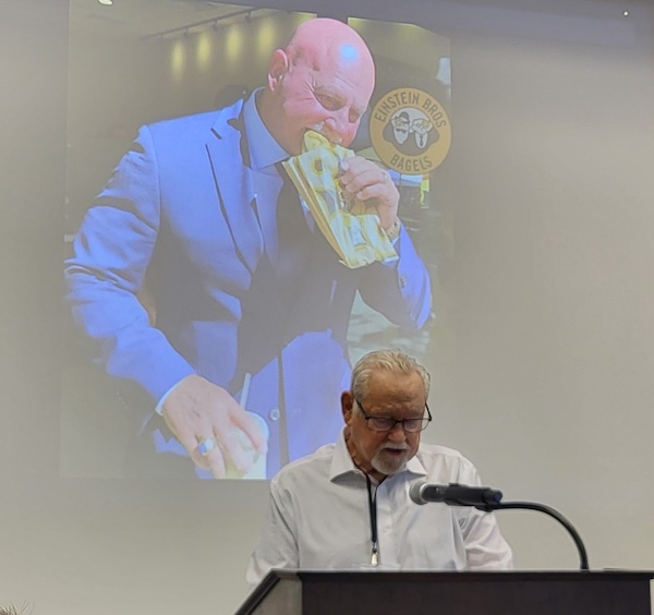 man at a podium with a photo of a man eating a bagel projected behind him