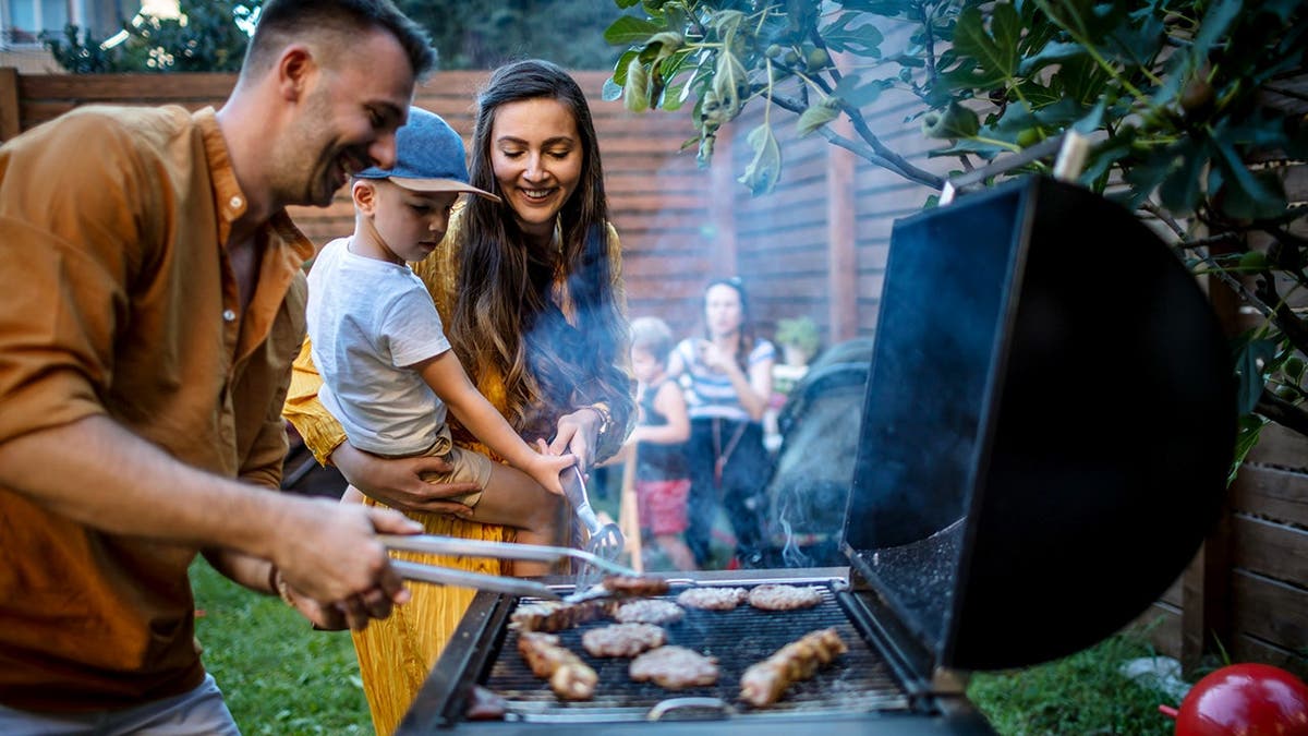 A young couple with a child smile while they grill meats on a backyard grill.