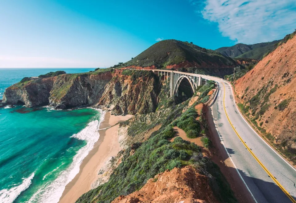 The Pacific coastline from Highway 1 in California shows a sweeping road and bridge alongside cliffs, a beach, and the ocean. Markus – stock.adobe.com