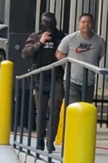 A law enforcement officer in dark clothing escorts a man in a gray Nike shirt along a walkway with yellow bollards.